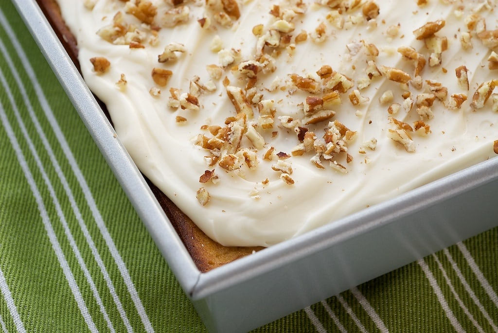 Overhead view of the corner of a banana cake in a metal baking pan, topped with frosting and chopped nuts