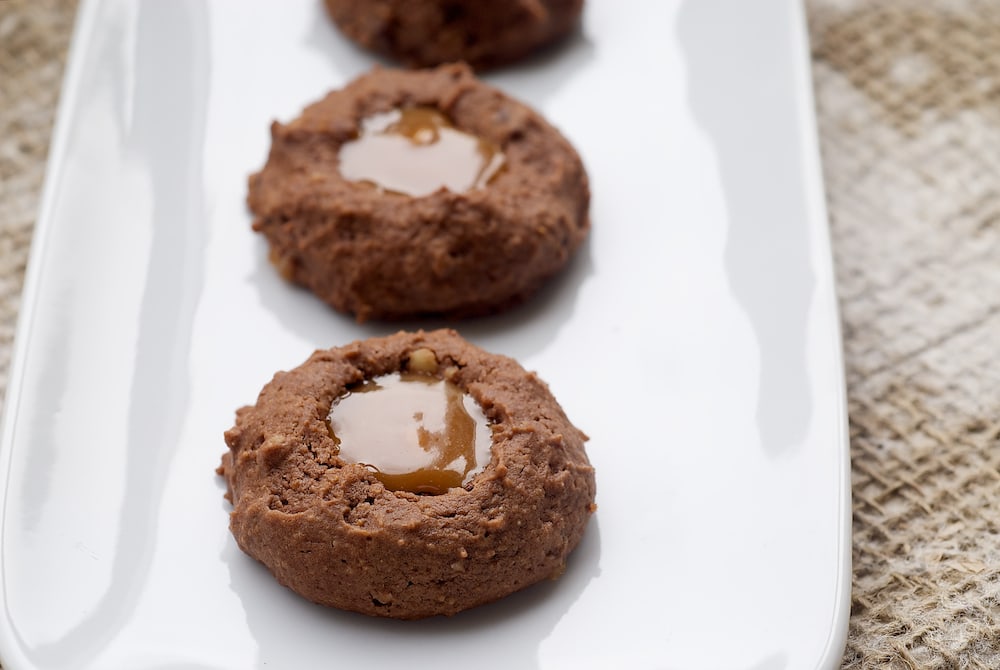 Turtle cheesecake thumbprint cookies in a row on a plate.