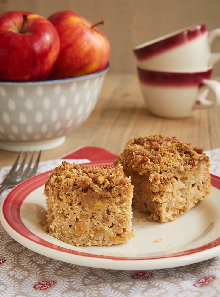 Two squares of Caramel Apple Crumb cake on plate with apples and teacups in background