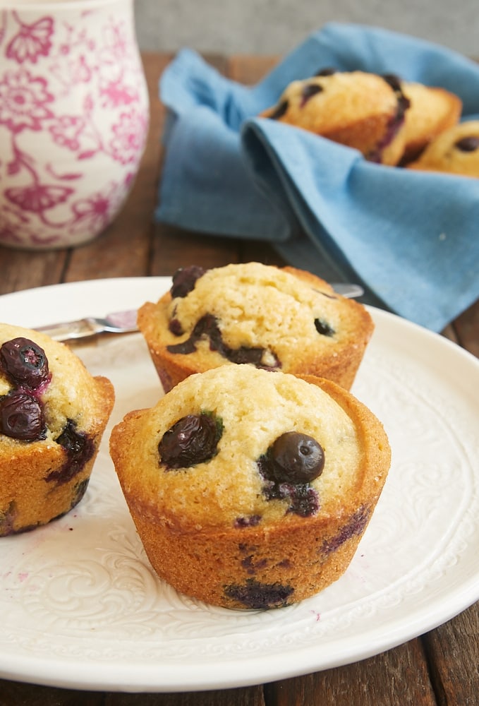 three blueberry muffins on a white plate with more muffins in a basket in the background
