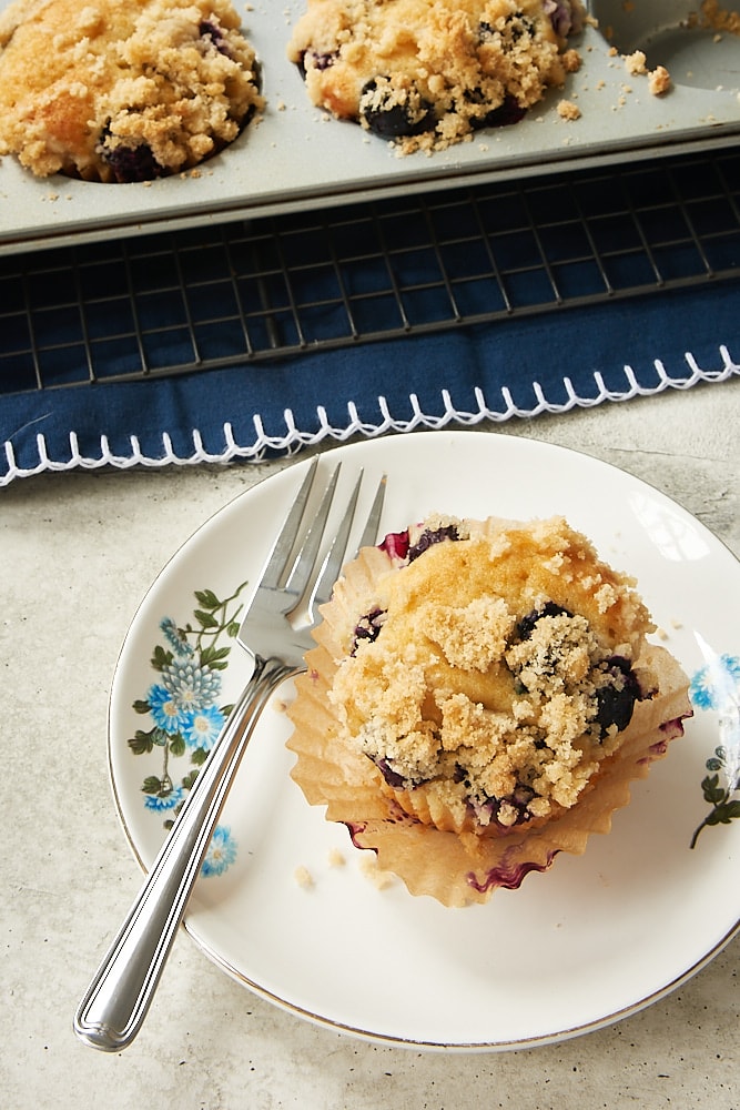 Blueberry Coffee Cake Muffin on a blue floral-edged plate with a partial view of more muffins in a muffin pan in the background