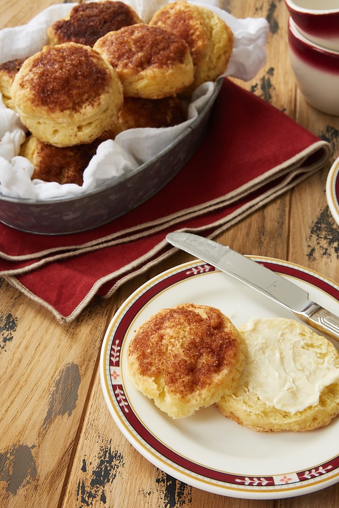 a split, buttered Cinnamon Sugar Biscuit on a red-rimmed plate with a basket of biscuits in the background