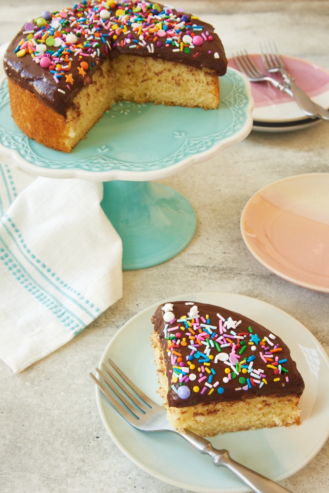 Slice of mini yellow cake with chocolate frosting on plate, with remaining cake on stand