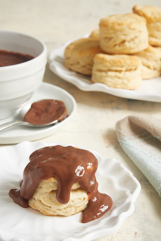 Biscuit topped with chocolate gravy on white plate, with additional biscuits in background