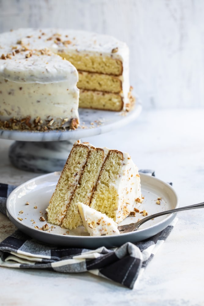 slice of Butter Pecan Cake on a beige plate with the remaining cake on a marble cake stand