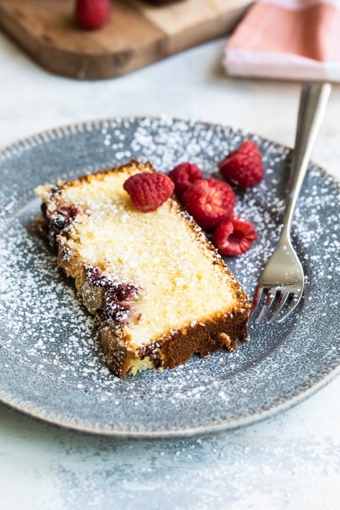 Slice of almond loaf dusted with powdered sugar, on plate with raspberries and fork