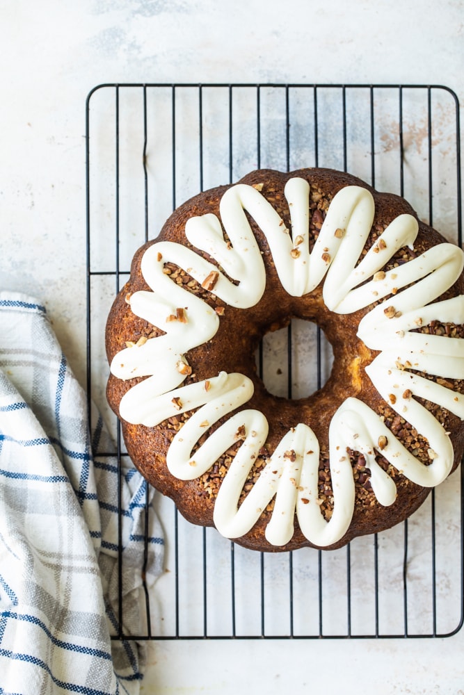 Whole hummingbird Bundt cake on wire rack with cream cheese glaze and pecans