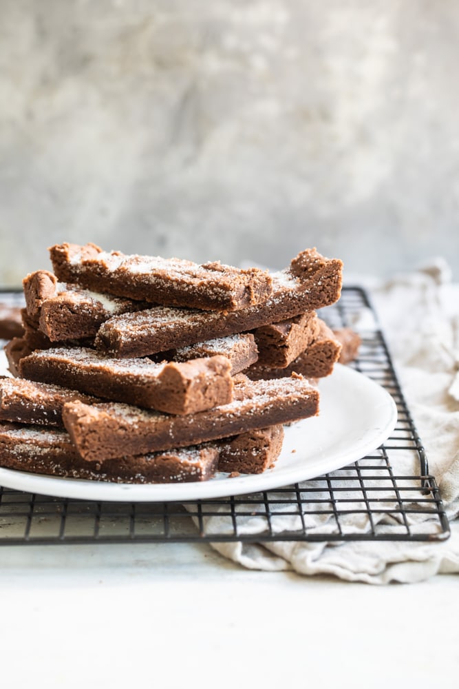 Chocolate shortbread cookies stacked on a plate set on a cooling rack.