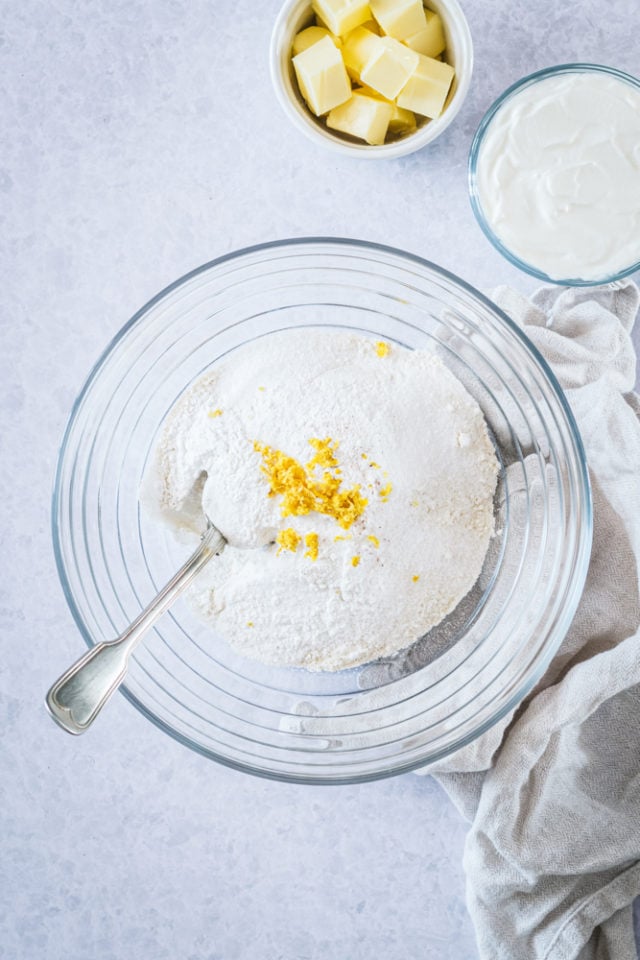 Overhead view of sugar, flour, baking powder, lemon zest, and salt in a glass mixing bowl.