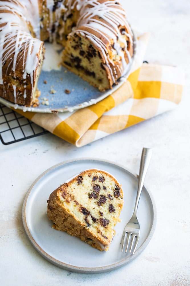 Slice of chocolate chip almond cake on plate with fork, with rest of cake in background