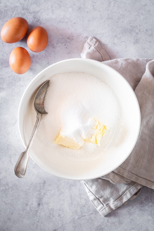 Overhead shot of sugar and butter in large white bowl with spoon, with whole eggs in background