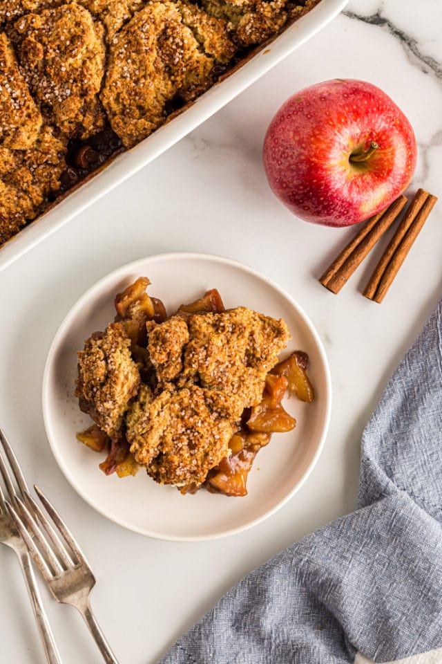 overhead view of Brown Sugar Apple Cobbler on a white plate with an apple, cinnamon sticks, and the remaining cobbler surrounding