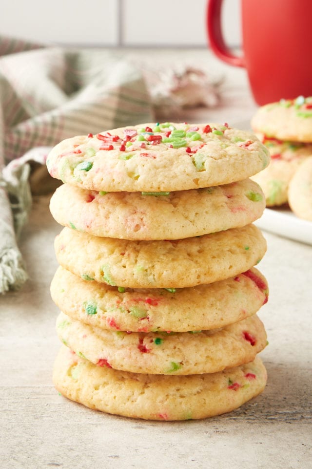 stack of Soft Christmas Sprinkle Sugar Cookies with more cookies and a red mug behind