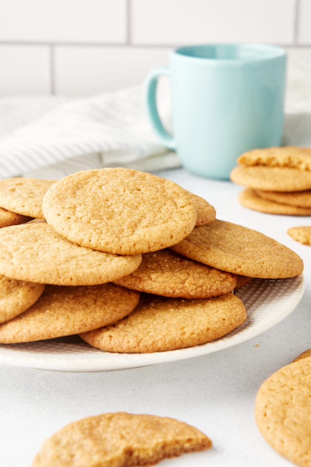 Maple Ginger Cookies piled on a white plate with more cookies and a blue-green mug
