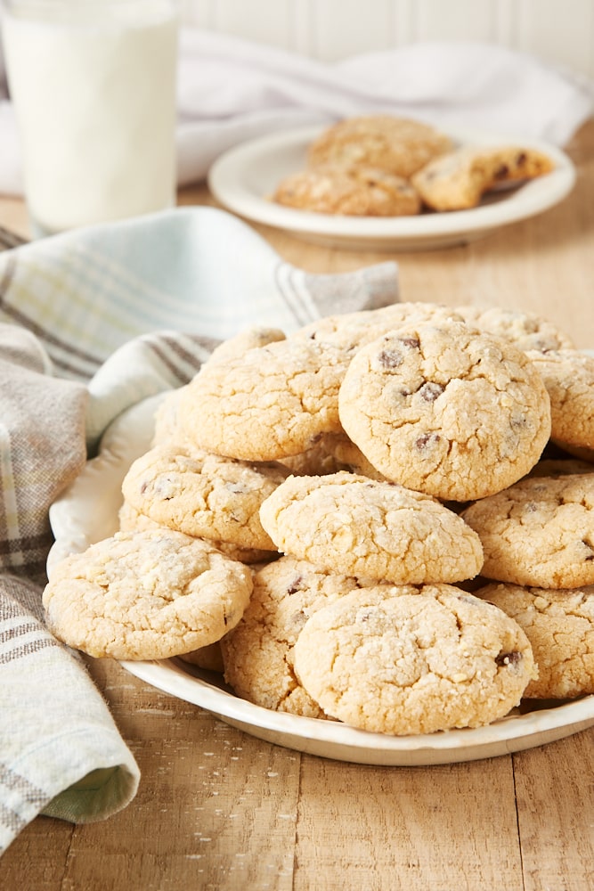 Pile of peanut butter crinkle cookies on a white plate with more cookies and a glass of milk in the background.