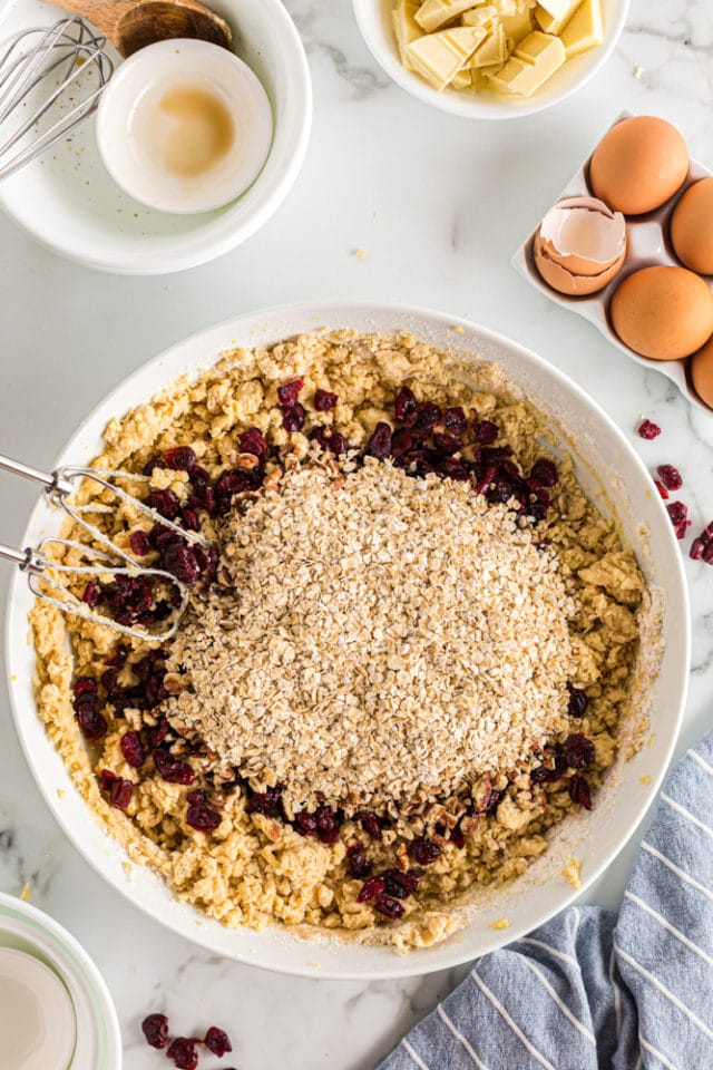 Overhead view of cranberries, pecans, and oats added to cookie dough in a white mixing bowl.