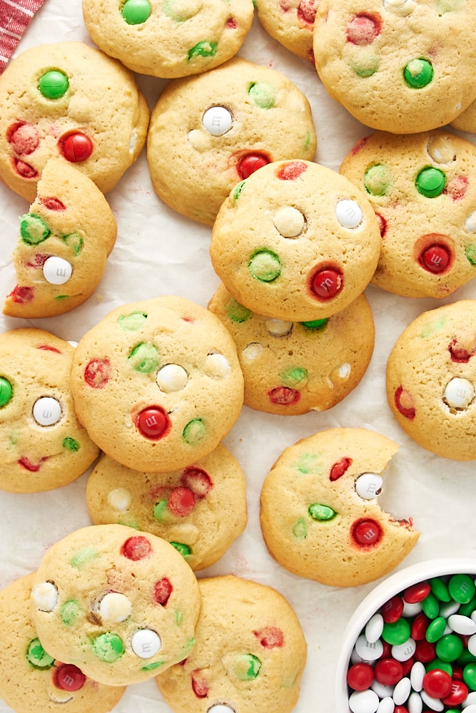Overhead view of Christmas M&M cookies scattered and piled on white parchment paper.