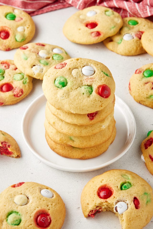 Christmas M&M Cookies stacked on a white plate with more cookies surrounding