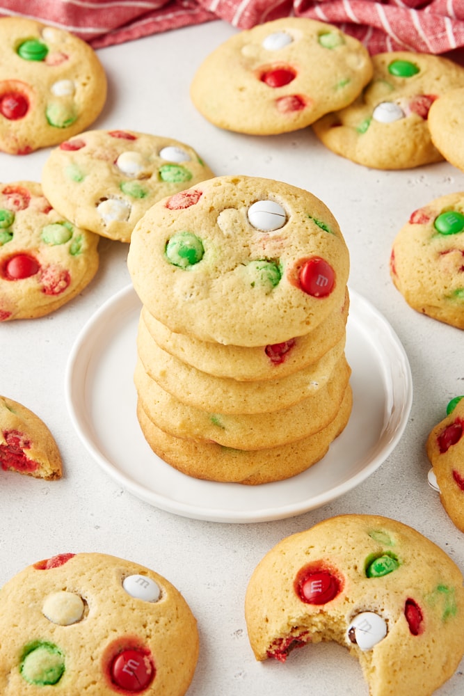 M&M cookies stacked on a white plate with more cookies surrounding.