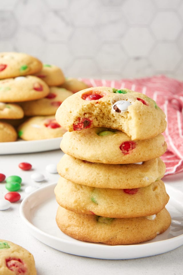 five Christmas M&M Cookies stacked on a white plate with a bite missing from the top one