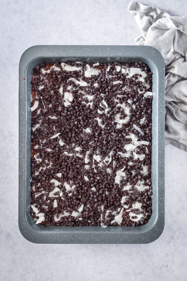 overhead view of coconut and chocolate chips sprinkled over brownie batter in a rectangular baking pan