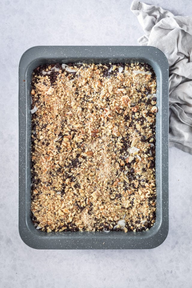 overhead view of coconut, chocolate chips, and chopped pecans sprinkled over brownie batter in a rectangular baking pan