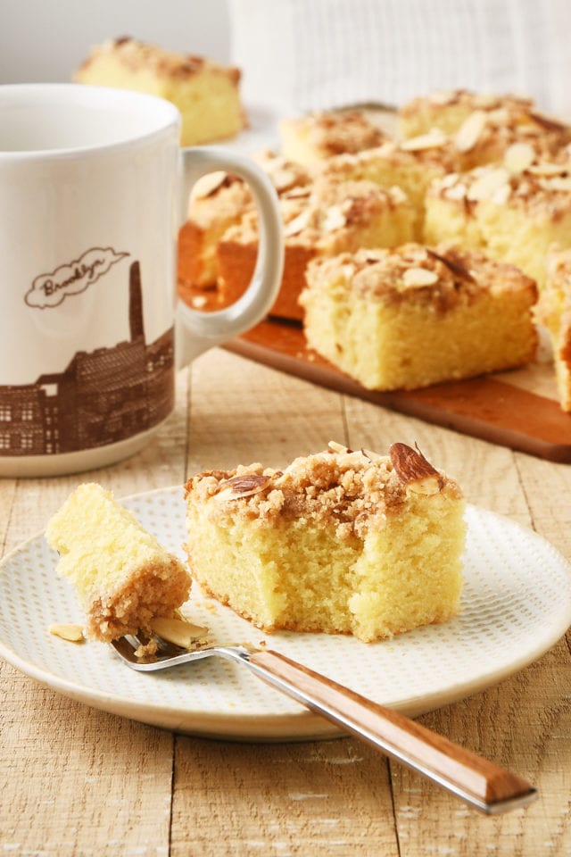 slice of Almond Crumb Cake with a bite on a fork on a white plate with a coffee cup and more cake in the background