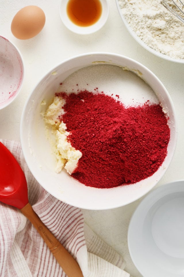Overhead view of sugar and freeze-dried raspberries added to a butter-cream cheese mixture in a white mixing bowl.