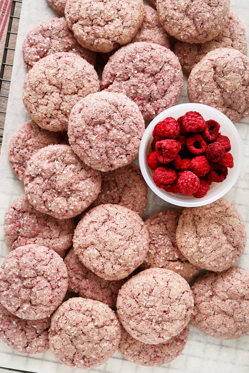 Overhead view of raspberry sugar cookies piled on parchment paper on a wire rack with a bowl of freeze-dried raspberries.