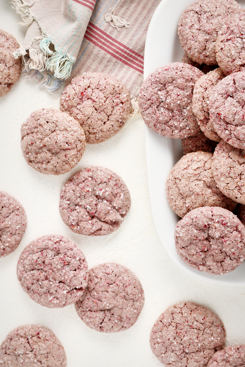 Overhead view of raspberry sugar cookies scattered on a white surface and piled on a white tray.