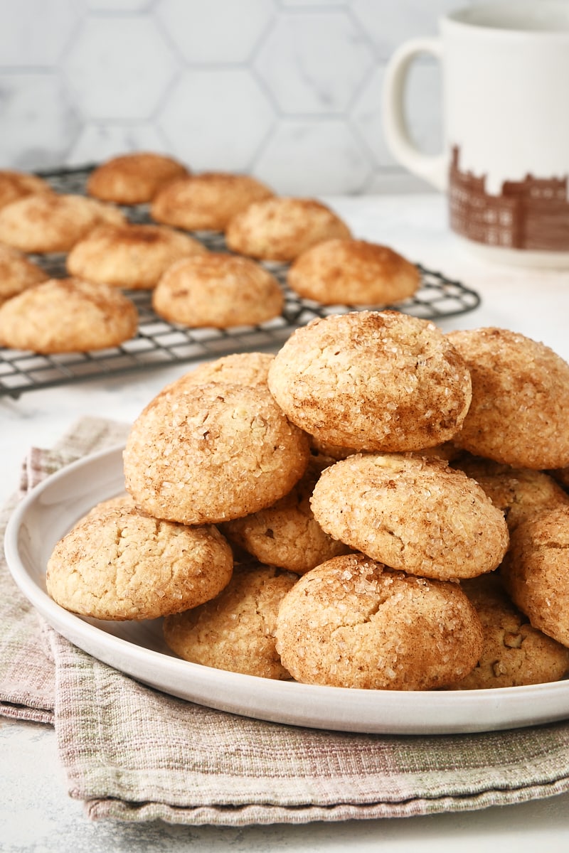 Brown Butter Cinnamon Cookies piled on a light gray plate with more cookies on a cooling rack in the background