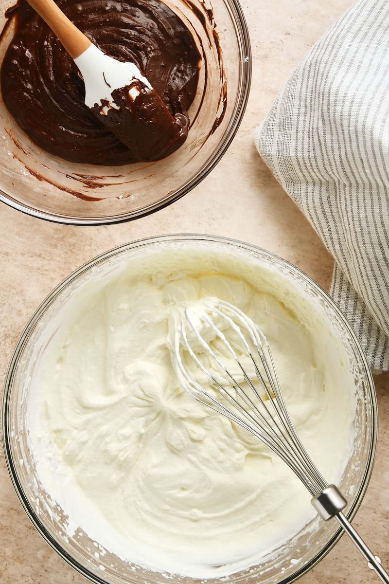 overhead view of whipped cream in a glass mixing bowl and melted chocolate mixture in another glass mixing bowl