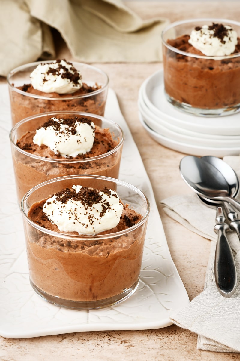 3 servings of Chocolate Mousse in clear glasses on a long white tray with another serving on a white plate in the background