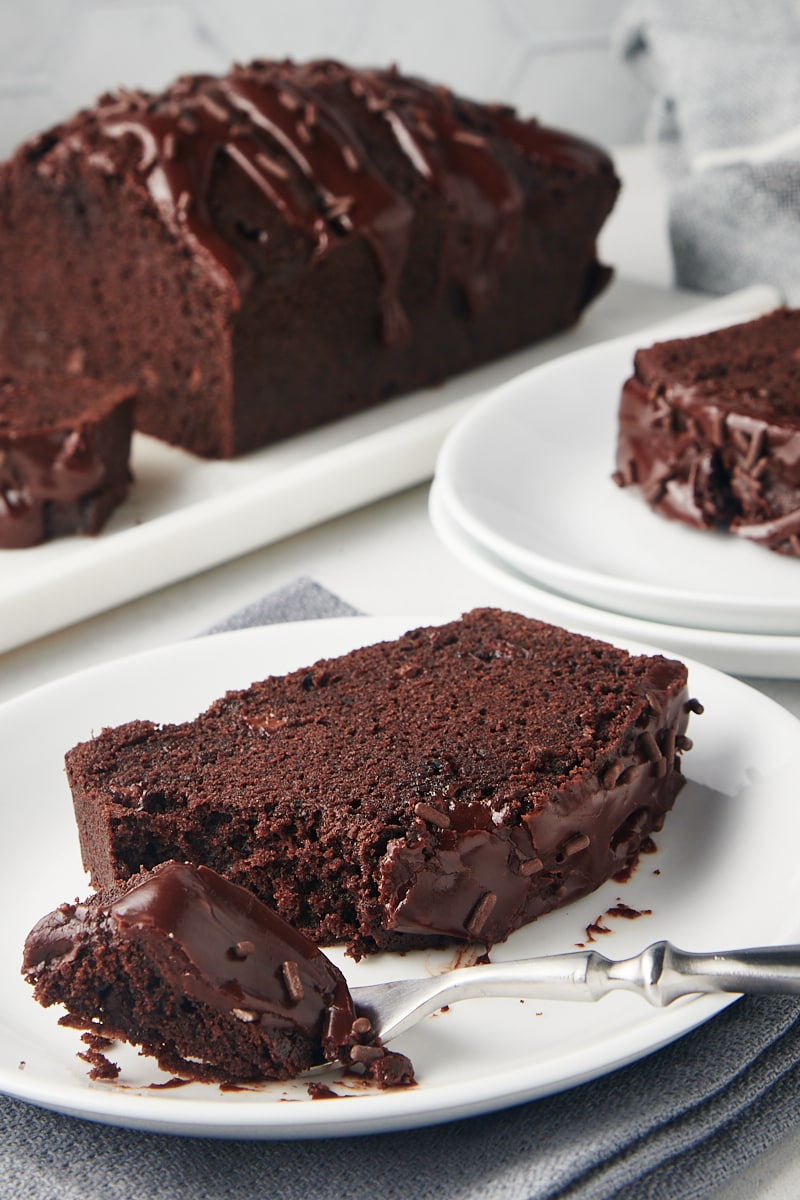 slice of Chocolate Loaf Cake on a white plate with more cake in the background