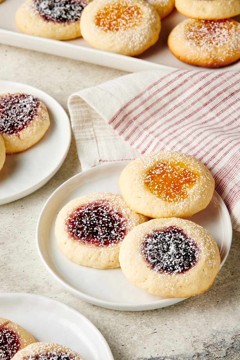 three Jam-Filled Cream Cheese Thumbprint Cookies on a white plate with more cookies in the background