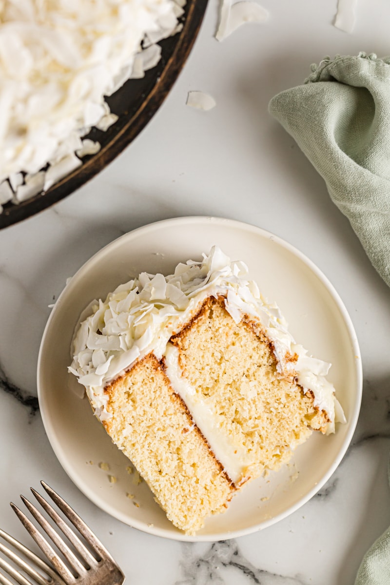 Slice of coconut cake on plate with rest of cake in background