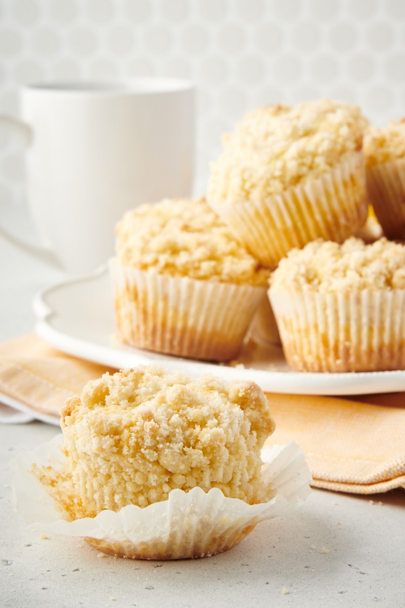 a partial Lemon Crumb Muffin on a white surface with more muffins on a plate behind