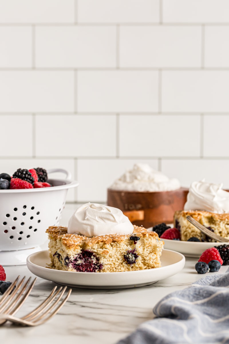 Square of berry vanilla cake on plate with dollop of whipped cream, berries, whipped cream, and another plate in background