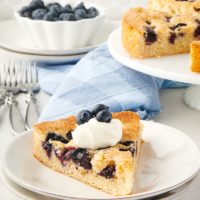 Slice of blueberry cake on plate with whipped cream and blueberries