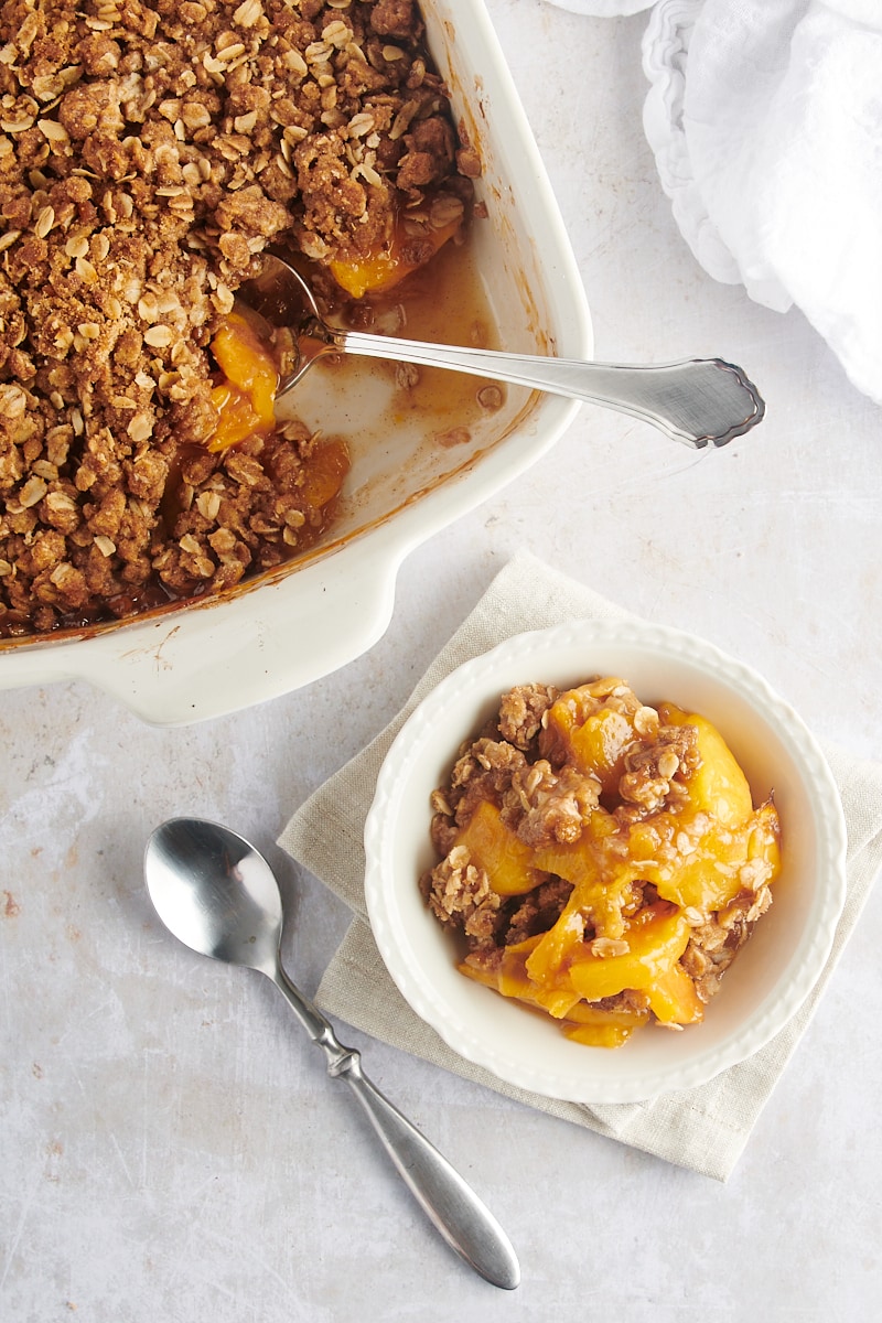 overhead view of a serving of peach crisp in a white bowl with remaining crisp in a baking pan