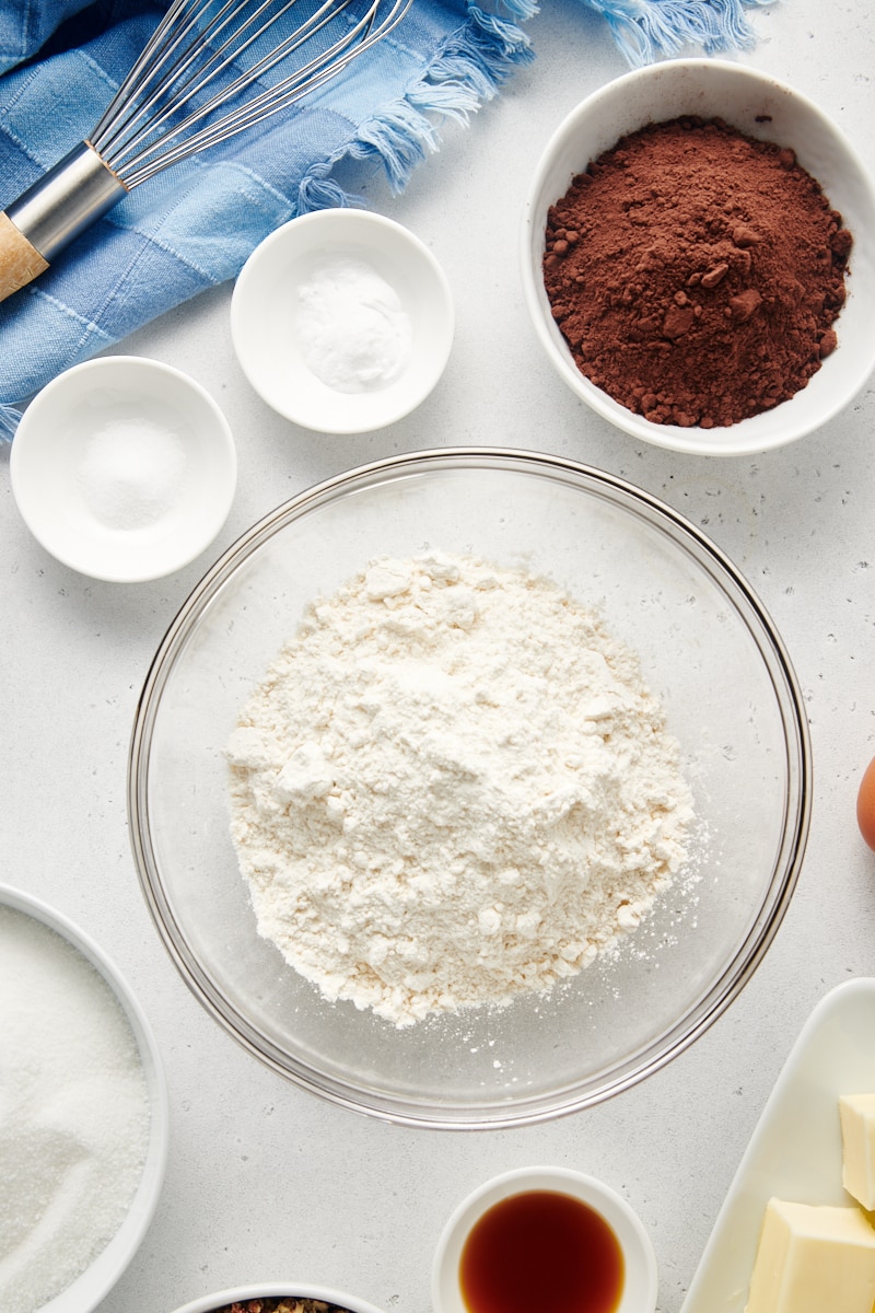 Flour in glass mixing bowl with other dry ingredients in smaller bowls