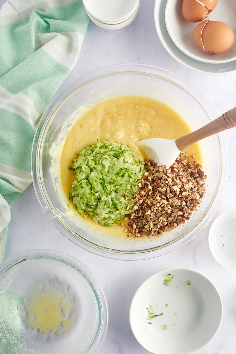 overhead view of zucchini and nuts added to muffin batter in a glass mixing bowl