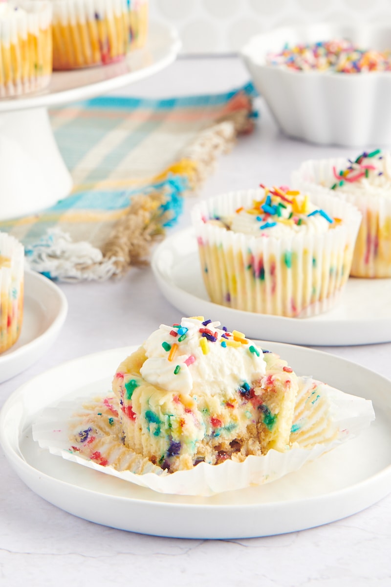 a Mini Funfetti Cheesecake on a white plate with more cheesecakes in the background. A bite is missing from the cheesecake.