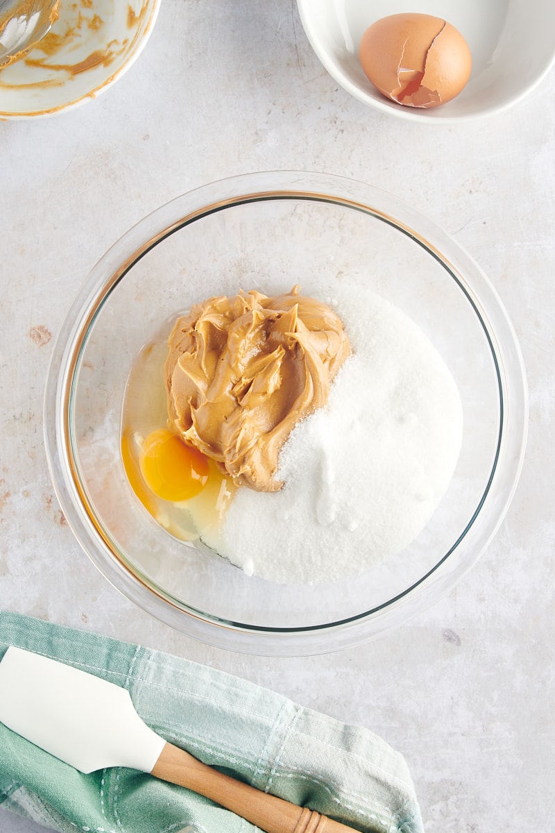 overhead view of peanut butter, sugar, and an egg in a glass mixing bowl