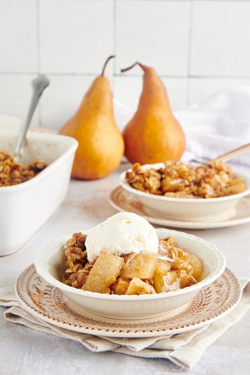 Brown Butter Pear Crisp with ice cream in a white bowl with more of the dessert and two pears in the background