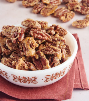 glazed pecans in a white bowl with more pecans scattered in the background