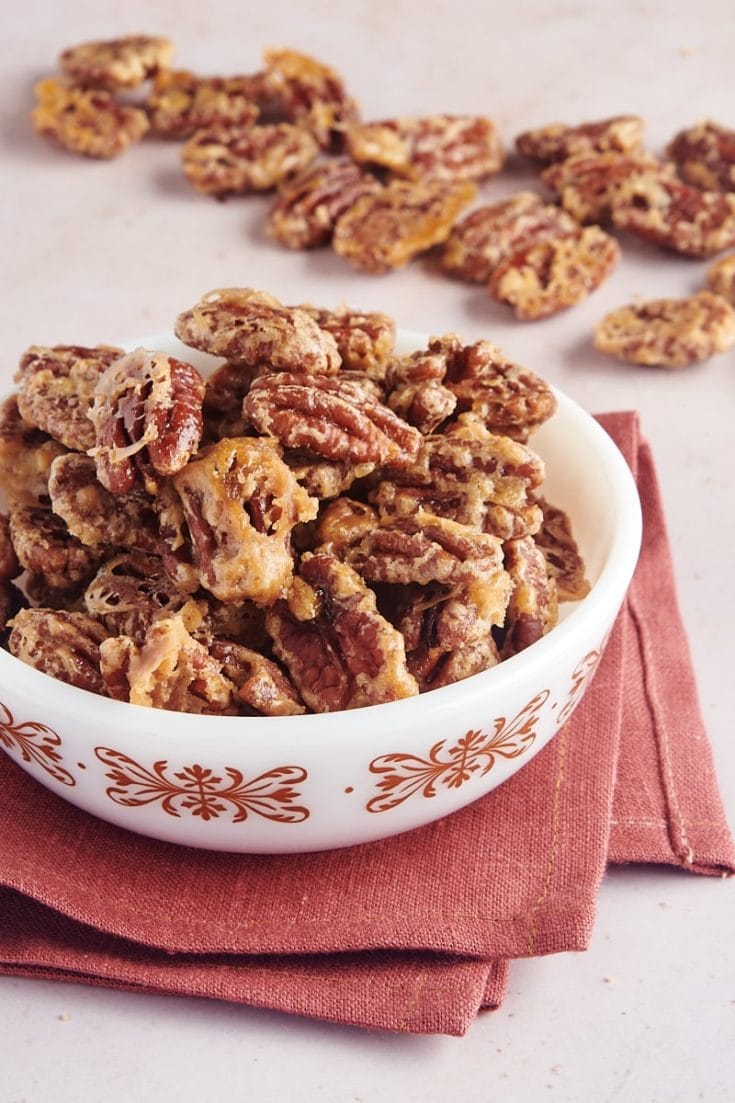 glazed pecans in a white bowl with more pecans scattered in the background