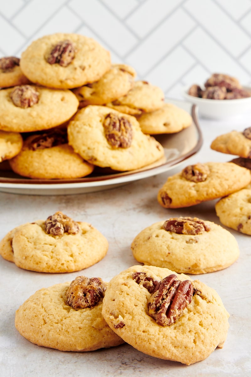 Maple Pecan Cookies spredt på en bordplade med flere cookies på en tallerken i baggrunden