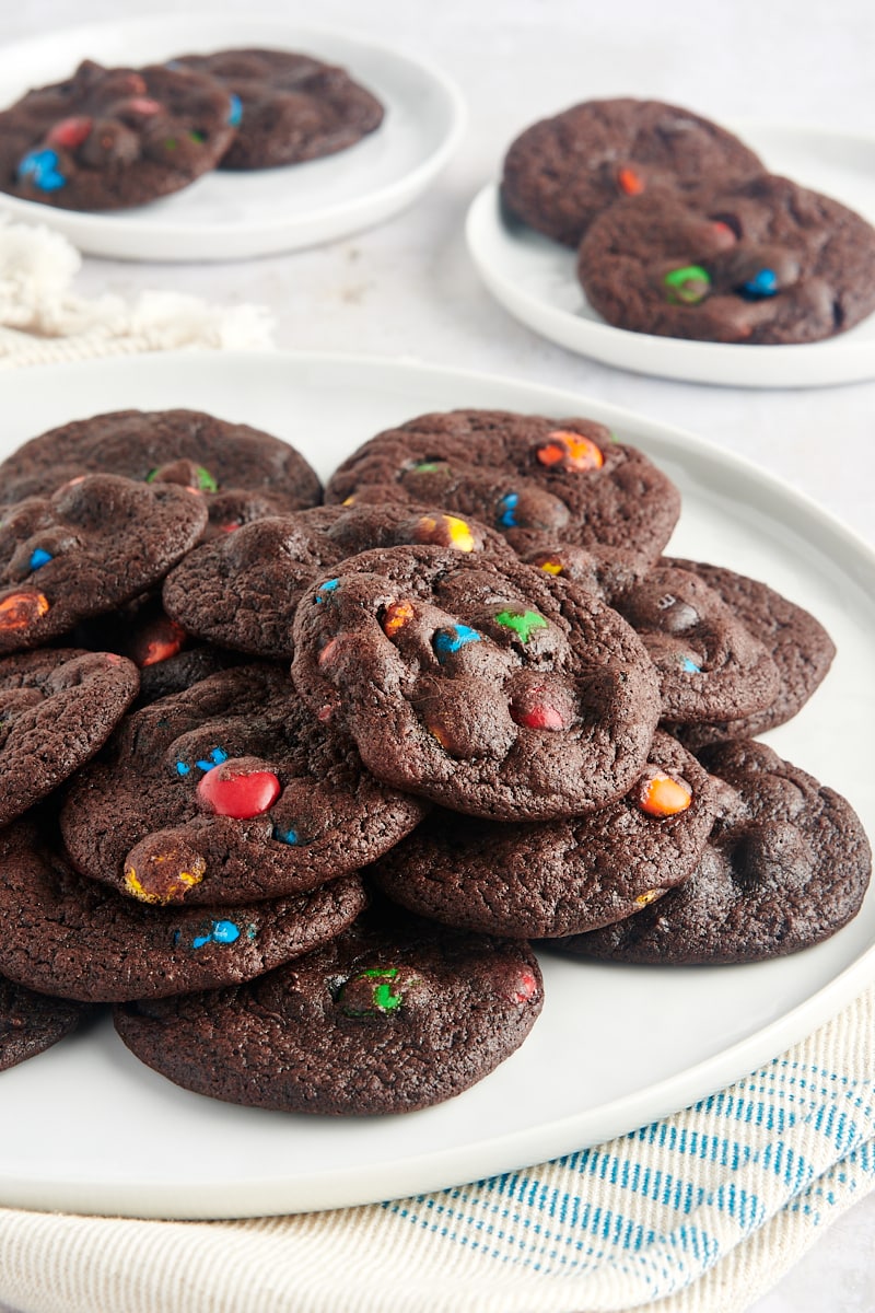 Chocolate M&M Cookies piled on a large white plate with more cookies on small plates in the background