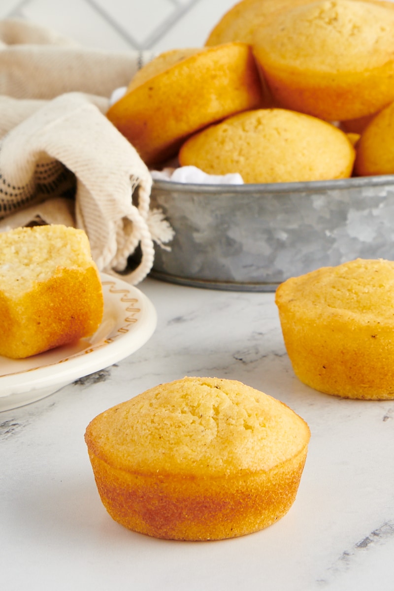 Corn muffins sitting on the counter with a basket of corn muffins in the background.